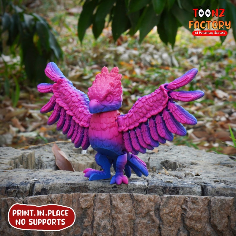 May include: A 3D-printed bird figurine with outstretched wings, featuring a gradient of pink, purple, and blue hues. The bird is perched on a tree stump. The text "PRINT.IN.PLACE NO SUPPORTS" is visible.