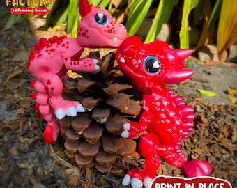 May include: Two 3D printed red and pink dragon figurines with large eyes and hearts on their cheeks. The dragons are posed on a pine cone. The text "PRINT.IN.PLACE NO SUPPORTS" is printed on the bottom of the image.