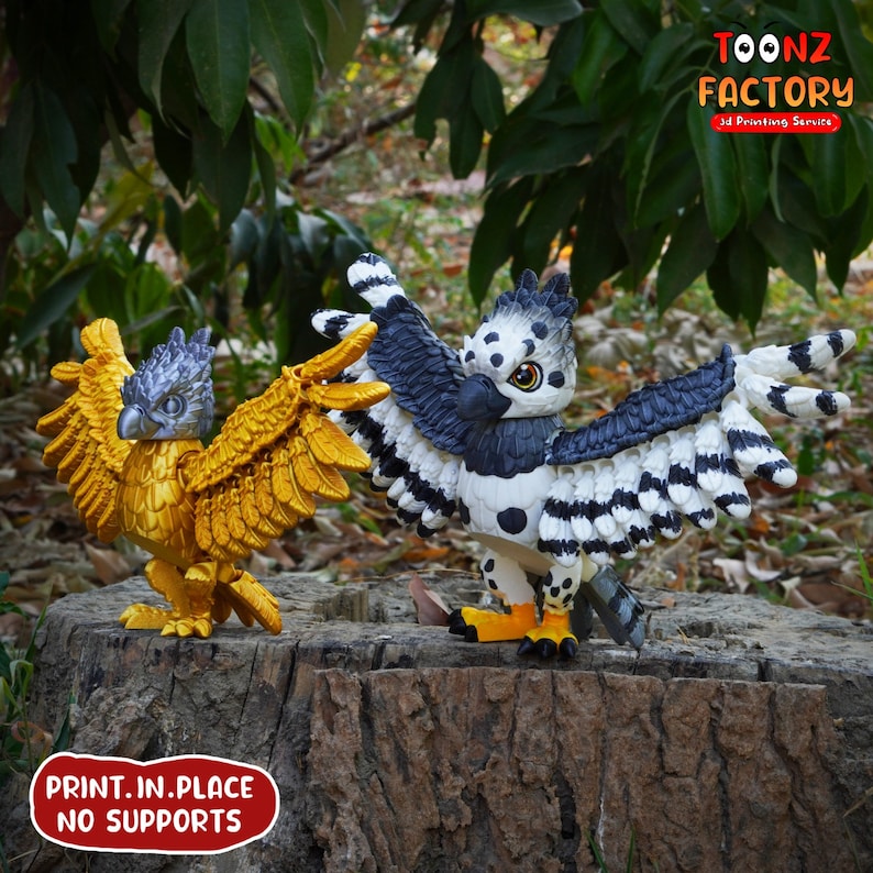May include: Two 3D-printed griffin figurines. One is gold, the other is black, white, and gray. The griffins are perched on a tree stump. The text "TOONZ FACTORY 3D Printing Service" and "PRINT.IN.PLACE NO SUPPORTS" are visible.