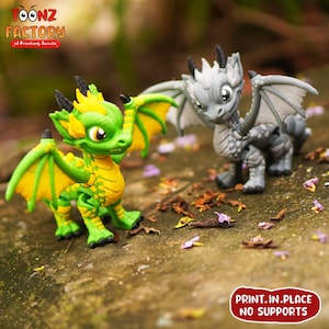K&ouml;nnte beinhalten: Zwei 3D-gedruckte Drachenfiguren, eine gr&uuml;n und gelb, die andere grau. Beide Drachen haben Fl&uuml;gel und stehen auf einer Holzoberfl&auml;che. Der Text "PRINT. IN. PLACE NO SUPPORTS" ist im Bild sichtbar.