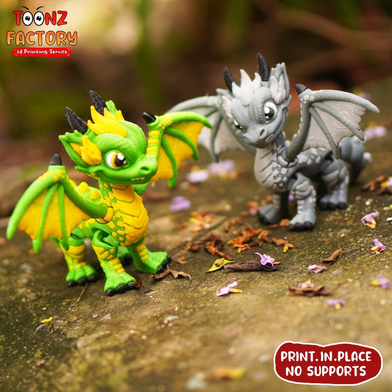K&ouml;nnte beinhalten: Zwei 3D-gedruckte Drachenfiguren, eine gr&uuml;n und gelb, die andere grau. Beide Drachen haben Fl&uuml;gel und stehen auf einer Holzoberfl&auml;che. Der Text "PRINT. IN. PLACE NO SUPPORTS" ist im Bild sichtbar.
