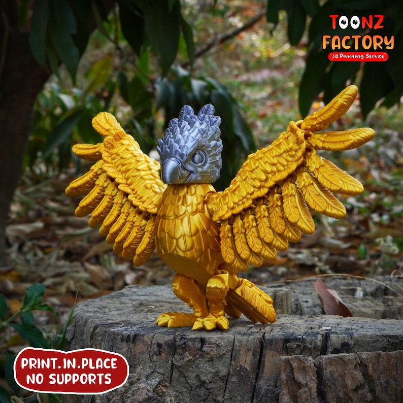 May include: A golden griffin figurine with outstretched wings, a silver head, and detailed feather textures. The figurine is 3D printed and stands on a tree stump. The image includes the text "TOONZ FACTORY 3D Printing Service" and "PRINT.IN.PLACE NO SUPPORTS".