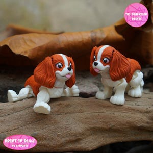 Puede incluir: Dos figuras de Cavalier King Charles Spaniel. Los perros son blancos, marrones y naranjas, con la lengua fuera. Las figuras están sobre un trozo de madera. La imagen incluye el texto "TRI DIMENSION PRINTS" y "PRINT IN PLACE NO SUPPORTS".