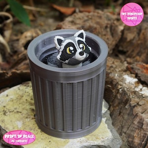 May include: A grey 3D-printed bin with a raccoon figurine inside. The raccoon has black and white fur with yellow eyes. The bin has vertical lines and is sitting on a rock. The text "PRINT IN PLACE NO SUPPORTS" is visible.