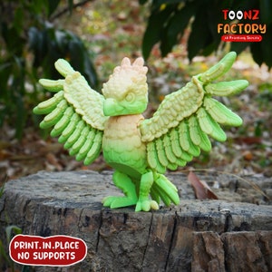 May include: A 3D-printed, articulated bird figurine with a gradient color scheme of green, yellow, and peach. The wings are spread, showcasing detailed feather textures. The text "PRINT.IN.PLACE NO SUPPORTS" is visible. The Toonz Factory logo is in the upper right corner.