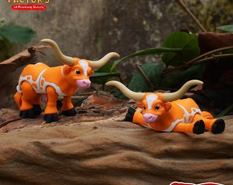 May include: Two orange and white 3D printed longhorn steer figurines. The figurines are posed on a brown wooden surface. The text "PRINT.IN.PLACE NO SUPPORTS" is printed on a red banner in the lower right corner of the image.