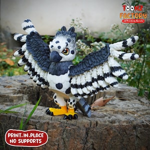 May include: A 3D-printed figurine of a stylized bird with outstretched wings. The bird features a black, white, and gray color scheme with yellow feet and a detailed design. The text "PRINT.IN.PLACE NO SUPPORTS" is visible.