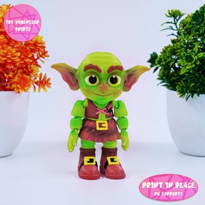 May include: A green goblin figurine with large ears, a red and brown outfit, and a pink cross on its chest. The figurine is standing on a white surface. The text "PRINT IN PLACE NO SUPPORTS" is visible in the lower right corner of the image.