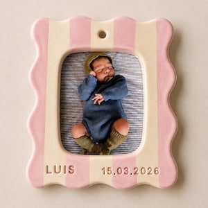 Pode incluir: Uma moldura retangular de cerâmica com borda ondulada, apresentando listras verticais em rosa claro e creme. A moldura exibe uma fotografia de um bebê e inclui o texto em relevo LUIS e 15.03.2026 na parte inferior, com um orifício no topo.