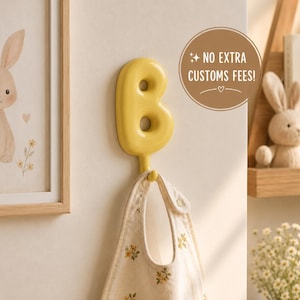 Op de afbeelding: Een glanzende gele wandhaak in de vorm van de hoofdletter B. De haak is aan een muur bevestigd en houdt een beige stoffen slabbetje met een klein geel bloemenpatroon vast. Een cirkelvormige grafische overlay op de afbeelding bevat de tekst NO EXTRA CUSTOMS FEES.