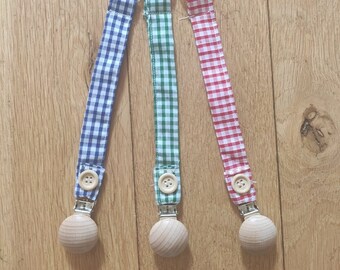May include: Three baby pacifier clips with gingham fabric straps in blue, green, and red. Each clip has a wooden button and a wooden ball-shaped end. The clips are on a light brown wooden surface.