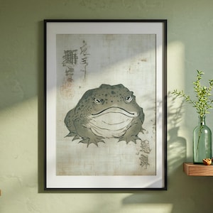 Puede incluir: Impresión enmarcada de un sapo de aspecto gruñón en tonos grises y verdes, inspirada en el arte japonés. La obra de arte presenta caracteres japoneses sobre un fondo texturizado blanquecino. La impresión está en un marco negro.