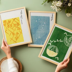 May include: Three rectangular framed prints with light wood frames. The left print features a yellow botanical pattern, the center print shows a blue textured landscape, and the right print displays a green background with white circular motifs.