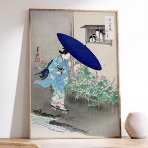 Puede incluir: Grabado japonés enmarcado que representa a una mujer con un kimono azul sosteniendo un gran paraguas azul. La obra de arte presenta un estilo tradicional con una paleta de colores apagados, mostrando una escena con un edificio y follaje.
