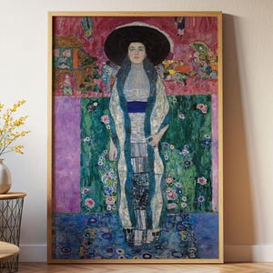 May include: A framed art print featuring Gustav Klimt's Lady with Hat and Feather Boa. The painting depicts a central figure in a high-collared white dress, a blue sash, and a large black hat, set against a vibrant background of pink, green, and blue.