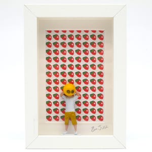 May include: Framed artwork showing a figure in a yellow mask and trousers against a strawberry backdrop. The white framed piece is signed in the bottom right corner. The artwork is in a white frame.