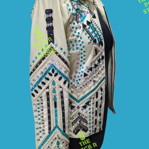 May include: A white leather jacket with turquoise and black geometric embroidery and rhinestones. The jacket has a zipper closure and long sleeves.