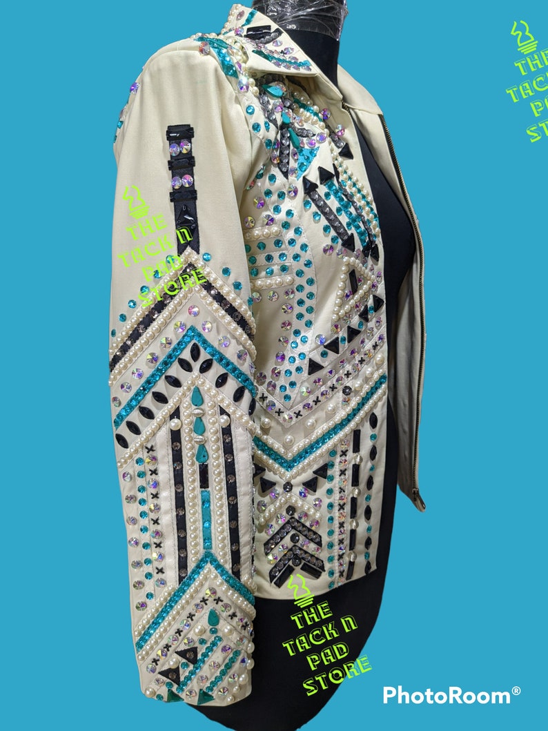 May include: A white leather jacket with turquoise and black geometric embroidery and rhinestones. The jacket has a zipper closure and long sleeves.