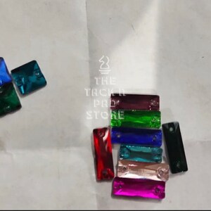 May include: Assortment of colorful glass rhinestones in square and rectangular shapes. The rhinestones are arranged in two groups on a white surface.