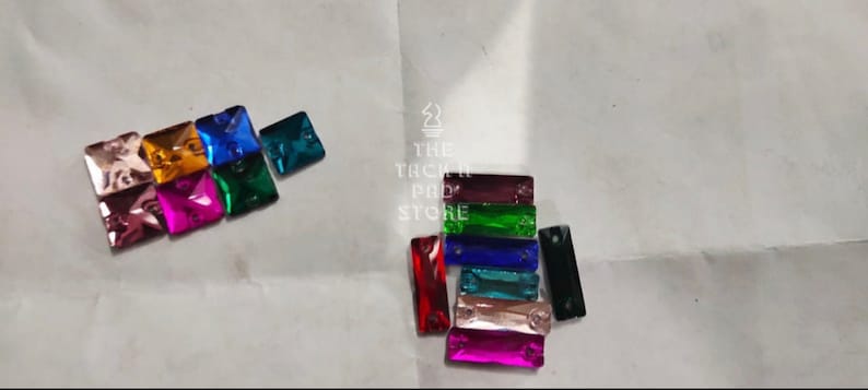 May include: Assortment of colorful glass rhinestones in square and rectangular shapes. The rhinestones are arranged in two groups on a white surface.