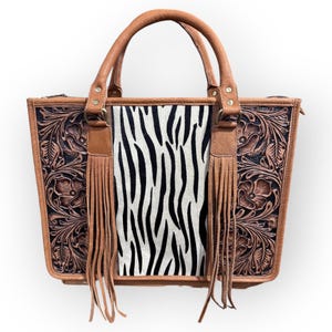 May include: A brown leather tote bag with a zebra print center panel. The bag features floral tooling on the sides, brown leather handles, and fringe accents. The bag has a rectangular shape.