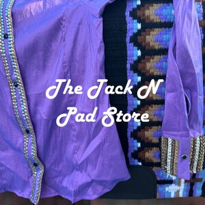 May include: A bright purple shirt with gold and silver detailing, alongside a patterned saddle blanket. The shirt has a button-down style and long sleeves. The text "The Jack N Pad Store" is displayed on the shirt.