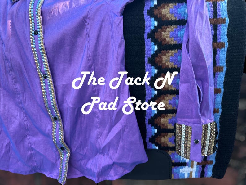 May include: A bright purple shirt with gold and silver detailing, alongside a patterned saddle blanket. The shirt has a button-down style and long sleeves. The text "The Jack N Pad Store" is displayed on the shirt.