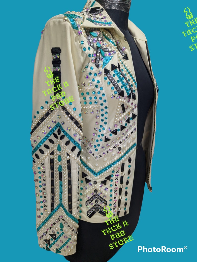 May include: A white leather jacket with turquoise and black geometric designs and pearl embellishments. The jacket has a zipper closure and a collar. The text "THE TACK N PAD STORE" is visible on the jacket.