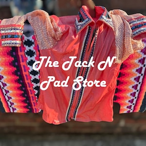 May include: Coral-colored western shirt with a collar and zipper, paired with a colorful saddle blanket. The blanket features a vibrant pattern of orange, pink, and purple. The text "The Jack N Pad Store" is visible.