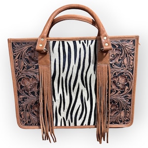 Puede incluir: Bolso tote de cuero marrón con un panel central con estampado de cebra. El bolso presenta un repujado floral en los laterales, un asa superior y flecos decorativos. El bolso mide aproximadamente 30 cm de ancho y 25 cm de alto.