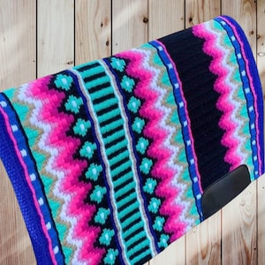 Può includere: Un'imbottitura per sella da cavallo dai colori vivaci. L'imbottitura presenta un pannello centrale nero, bordato da un motivo a zig-zag rosa, bianco e verde acqua. I bordi sono rifiniti in blu, con una toppa in pelle nera nell'angolo inferiore.