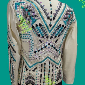 May include: A white show jacket with a turquoise, black, and white geometric design. The jacket is embellished with rhinestones and pearls. The text "The Tack Pad Store" is visible on the jacket.