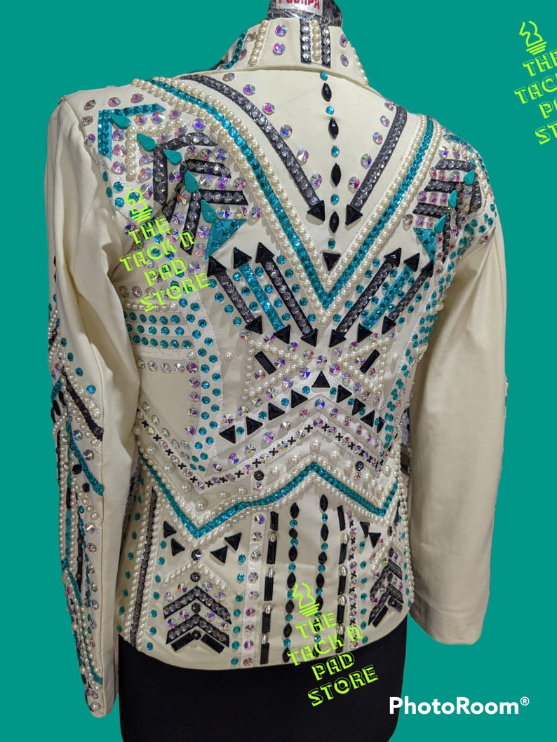 May include: A white show jacket with a turquoise, black, and white geometric design. The jacket is embellished with rhinestones and pearls. The text "The Tack Pad Store" is visible on the jacket.