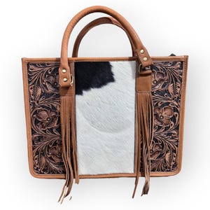 Puede incluir: Bolso tote rectangular con un panel central de piel de vaca y detalles en cuero marrón. El bolso presenta un diseño floral labrado en los laterales, asas de cuero y flecos decorativos. El bolso mide aproximadamente 30 cm de ancho y 25 cm de alto.