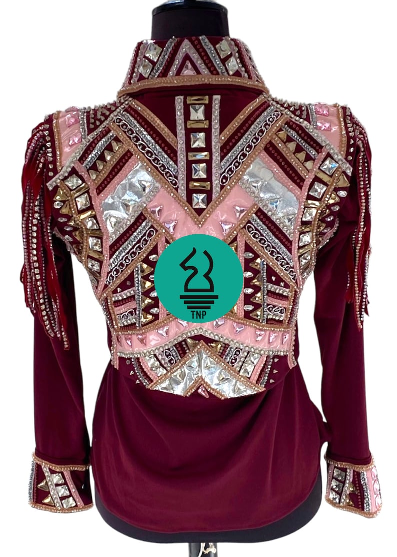 May include: Burgundy long-sleeve shirt with a pink, silver, and gold geometric design featuring rhinestones and fringe. The shirt has a collar and button closure. The back of the shirt features a logo that says "TNP".