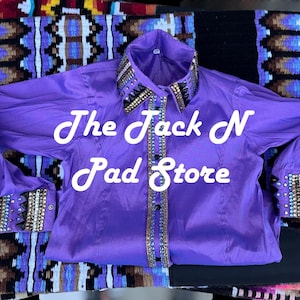 May include: A bright purple long-sleeved shirt with decorative detailing on the collar and cuffs. The shirt has the text "The Jack N Pad Store" in white script. It is displayed on a patterned textile.