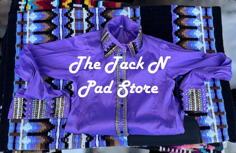May include: A bright purple long-sleeved shirt with decorative detailing on the collar and cuffs. The shirt has the text "The Jack N Pad Store" in white script. It is displayed on a patterned textile.