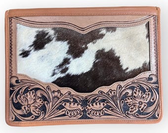 Puede incluir: Un bolso de mano rectangular de cuero con un panel central de piel de vaca. El bolso de mano presenta un borde marrón claro y un diseño floral decorativo en negro. La piel de vaca tiene un patrón en blanco y negro.