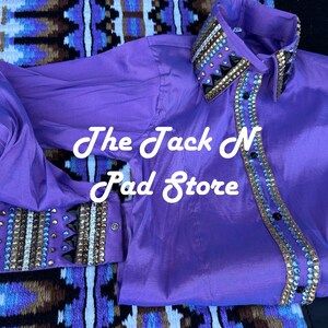 May include: A bright purple shirt with long sleeves, featuring decorative embellishments on the collar, cuffs, and front. The text "The Jack N Pad Store" is visible. The shirt is likely for equestrian use.