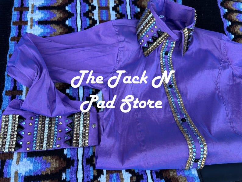 May include: A bright purple shirt with long sleeves, featuring decorative embellishments on the collar, cuffs, and front. The text "The Jack N Pad Store" is visible. The shirt is likely for equestrian use.