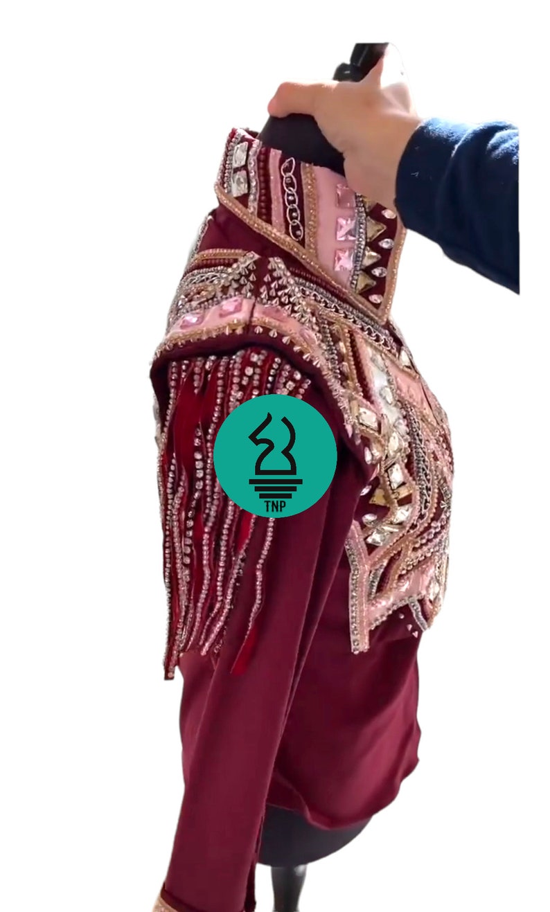 May include: A maroon show shirt with long sleeves and a beaded design. The shirt has a pink and gold trim with rhinestones and fringe. The shirt is on a mannequin.