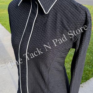May include: A black long-sleeve shirt with a textured honeycomb pattern, a pointed collar, and white piping trim along the front placket and collar edges. The text The Tack N Pad Store is visible across the front of the garment.