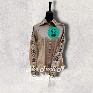 Könnte beinhalten: Ein langärmeliges, hellbraunes Hemd mit Knöpfen und einem geometrischen Muster an den Ärmeln. Das Muster besteht aus schwarzen, braunen und goldenen Perlen und Strasssteinen.