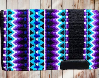 May include: A rectangular horse saddle pad with a black base and a vibrant pattern of turquoise, purple, and white geometric designs. The pad features a black leather patch at the bottom and is displayed on a wooden surface.