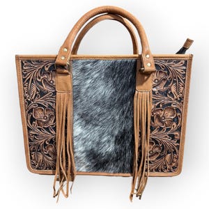 May include: A brown leather tote bag with a central panel of gray and white cowhide. The sides feature floral tooling on a black background, with brown leather handles and fringe.
