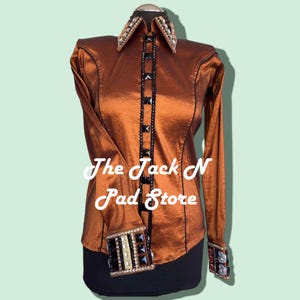 Puede incluir: Una camisa de manga larga de color óxido con adornos negros y dorados. La camisa tiene un cuello puntiagudo y cierre de botones en la parte delantera. El texto "The Jack N Pad Store" es visible en la camisa.