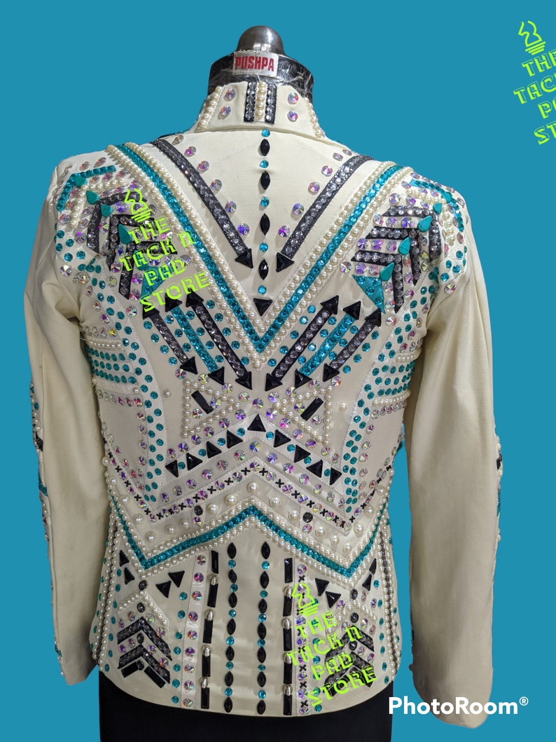 May include: A white show jacket with a geometric design featuring black, turquoise, and silver embellishments. The jacket has a collar and long sleeves. The text "The Tack Pad Store" is visible on the jacket.