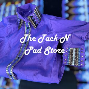 May include: A bright purple shirt with decorative beading on the collar and cuffs. The shirt has the text "The Jack N Pad Store" in white script. The fabric has a sheen, possibly satin or silk. The shirt is a striking colour.