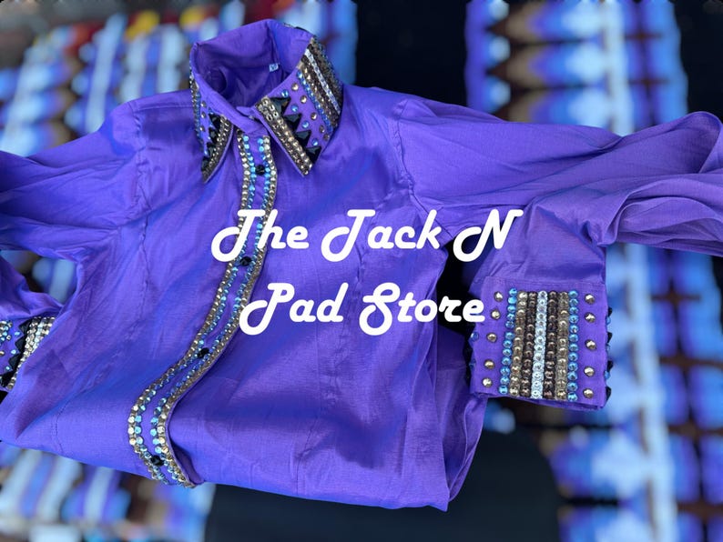 May include: A bright purple shirt with decorative beading on the collar and cuffs. The shirt has the text "The Jack N Pad Store" in white script. The fabric has a sheen, possibly satin or silk. The shirt is a striking colour.