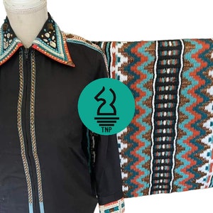 Könnte beinhalten: Schwarzes Reitturniershirt mit dekorativem Kragen, Manschetten und Schulterakzenten. Das Shirt hat einen durchgehenden Reißverschluss und wird mit einer gemusterten Satteldecke mit türkisfarbenen, orangefarbenen und weißen Zickzackmustern kombiniert.